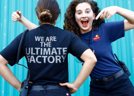 Smiling worker woman pointing at the t-shirt of another woman where it can be read: We are the ultimate factory